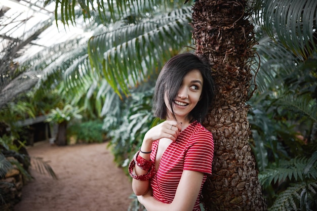 Plan intérieur d'une charmante jeune femme avec un joli sourire et des cheveux courts foncés, appuyée sur un grand palmier, regardant par-dessus son épaule et souriant de manière ludique. Concept de personnes, nature, style et mode