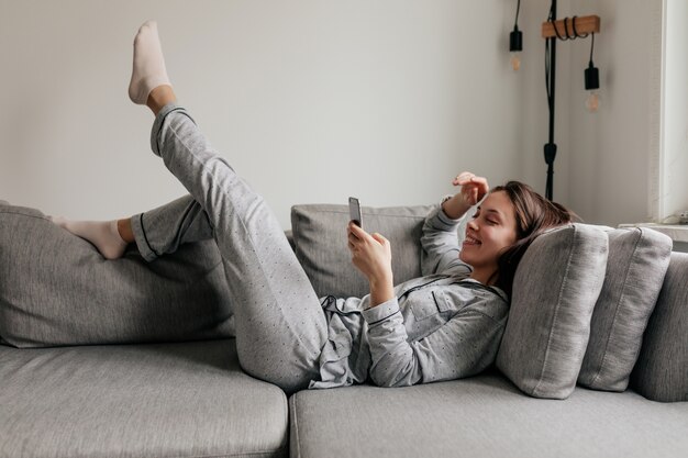 Plan intérieur complet d'une femme heureuse dans des vêtements à la maison faisant défiler le smartphone en position couchée sur l'entraîneur en journée ensoleillée.