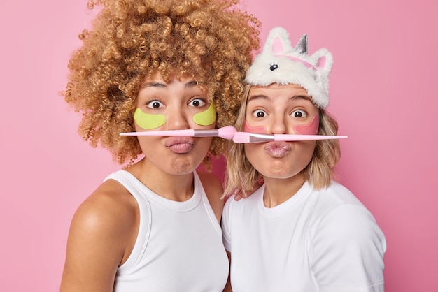 Photo gratuite plan intérieur de deux femmes surprises stupides tout en faisant des procédures de beauté boucher les lèvres tenir des pinceaux cosmétiques appliquer des patchs d'hydrogel pour réduire les rides porter des vêtements décontractés isolés sur rose