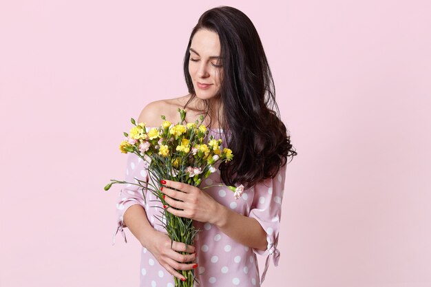 Plan intérieur du modèle féminin aux cheveux noirs heureux détient un bouquet de fleurs, vêtu d'une robe à la mode, isolé sur rose. Une jolie femme romantique reçoit des fleurs le 8 mars