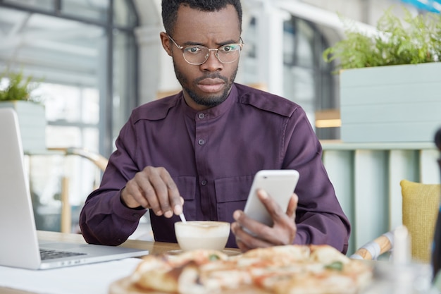 Plan intérieur d'un entrepreneur afro-américain surpris à la peau sombre reçoit de mauvaises nouvelles sur un téléphone intelligent, regarde l'écran, travaille sur un projet d'entreprise sur un ordinateur portable