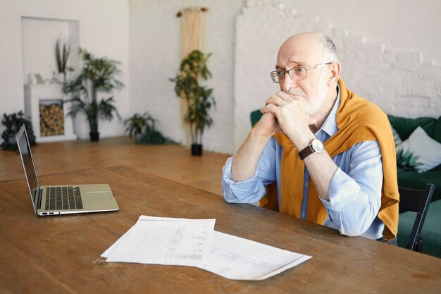 Plan intérieur d'un entrepreneur masculin barbu senior triste malheureux assis au bureau avec un ordinateur portable et des papiers ayant une expression faciale déprimée, frustré par des problèmes financiers, se tenant la main sous son menton