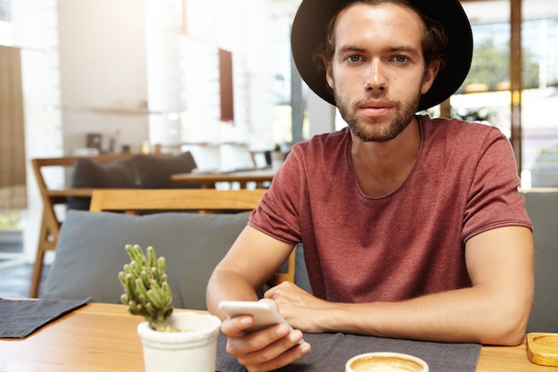 Plan intérieur d'un étudiant élégant au chapeau noir envoyant des SMS à des amis via les réseaux sociaux, en utilisant le Wi-Fi gratuit sur son téléphone portable pendant le petit-déjeuner au café avec un intérieur moderne, à la recherche