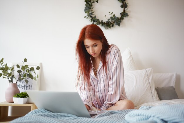 Plan intérieur d'une étudiante aux cheveux rouges assidue en vêtements de nuit élégants, assise sur un lit défait avec des écouteurs, utilisant une connexion Internet haute vitesse sans fil sur un ordinateur portable tout en faisant des travaux à domicile