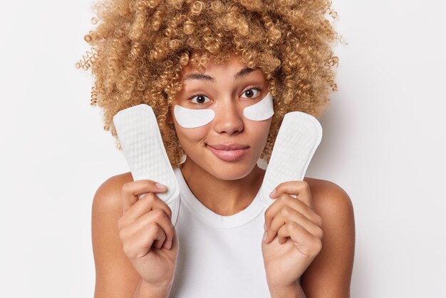 Plan intérieur d'une femme aux cheveux bouclés tenant des joints organiques respectueux de l'environnement applique des patchs de beauté sous les yeux pour éliminer les rides vêtue d'un t-shirt décontracté isolé sur fond blanc Hygiène féminine