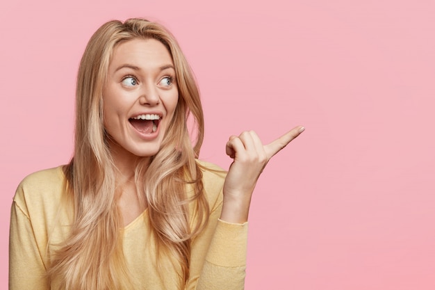 Photo gratuite plan intérieur d'une femme blonde étonnée a une expression ravie, indique avec un regard excité de côté, pose contre un mur rose avec un espace de copie vierge pour votre contenu publicitaire. wow, c'est génial!