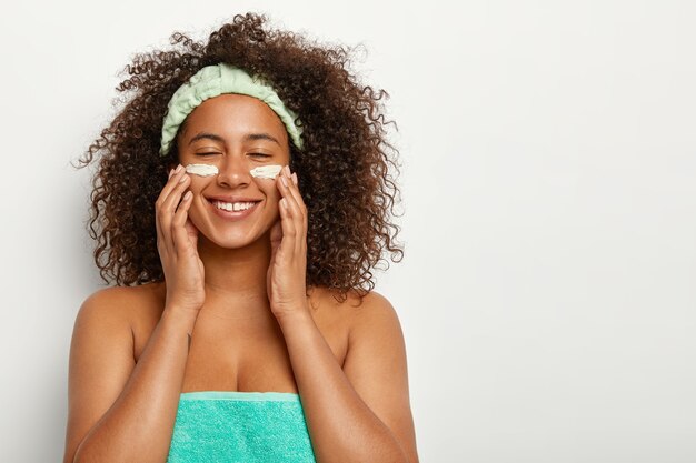 Plan intérieur d'une femme heureuse avec une coiffure afro, applique une crème cosmétique pour les soins de la peau, sourit positivement, a un visage frais et propre, utilise un hydratant de jour ou une lotion anti-âge, enveloppé dans une serviette turquoise