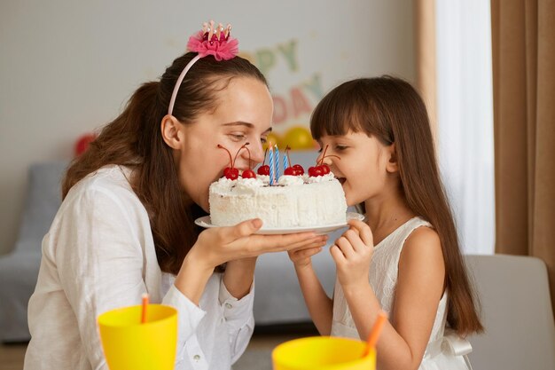 Plan intérieur d'une femme heureuse avec une petite fille assise à table et mordant un gâteau de dégustation, célébrant son anniversaire, exprimant des émotions festives positives, posant à l'intérieur à la maison.
