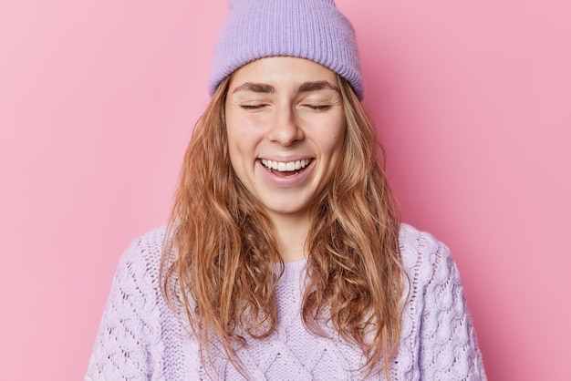 Plan intérieur d'une femme heureuse qui rêvasse et ferme les yeux a une mémoire heureuse rit positivement porte un chapeau et un pull tricoté sourit des poses heureuses sur fond rose. Notion d'émotions positives