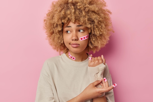 Photo gratuite plan intérieur d'une femme mécontente et malheureuse aux cheveux bouclés a une coupe au doigt applique un pansement adhésif porte un bandage sur le bras vêtu d'un t-shirt décontracté isolé sur fond rose la femme a un traumatisme