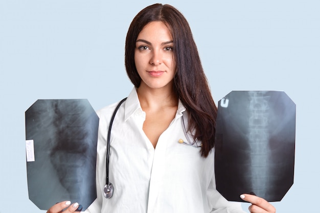 Plan intérieur d'une femme médecin brune sérieuse avec deux films radiographiques, examine la colonne vertébrale humaine, porte une robe blanche avec un stéthoscope, se tient dans la chambre du patient, isolé sur bleu clair.