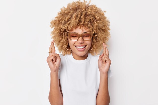 Plan intérieur d'une femme positive aux cheveux bouclés croise les doigts croit en la bonne chance espère que les rêves deviennent réalité porte des lunettes et un t-shirt décontracté isolé sur fond blanc Concept de langage corporel