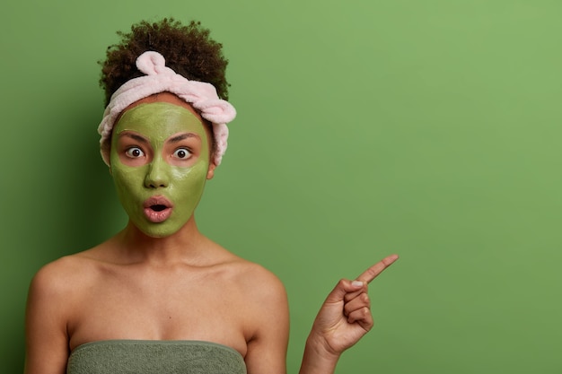 Plan intérieur d'une femme surprise émotionnelle fait des procédures de beauté, applique un masque facial pour le rajeunir, montre quelque chose de choquant sur un espace vide, un mur vert. Soins de la peau, concept de bien-être