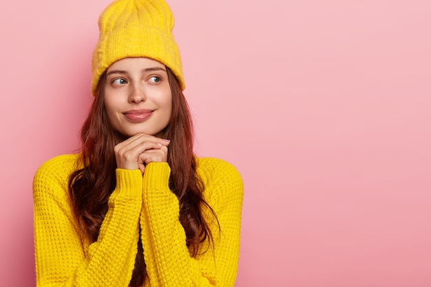 Plan intérieur d'une femme tendre et réfléchie garde les mains sous le menton, porte un chapeau jaune élégant et un pull chaud, pose contre un mur rose, un espace vide.