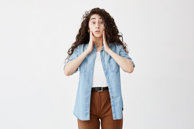 Plan Intérieur D'une Femme Terrifiée Portant De Longs Cheveux Ondulés Lâches, Vêtue D'une Chemise En Jean Et D'un Pantalon Brun, Garde Les Mains Sur Les Joues, Oublie De Faire Des Travaux. Fille Choquée Aux Yeux éclatés