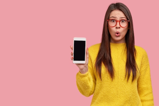 Plan intérieur d'une hipster brune surprise avec une expression étonnée montre un téléphone mobile avec écran blanc, voit des remises sur les gadgets en magasin, porte des vêtements jaunes