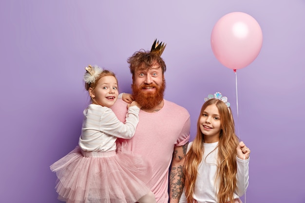 Photo gratuite plan intérieur d'un homme aux cheveux roux heureux avec une couronne sur la tête, porte une belle petite fille sur les mains, organise des vacances inoubliables pour sa fille lors de la journée internationale des enfants. famille de gingembre.