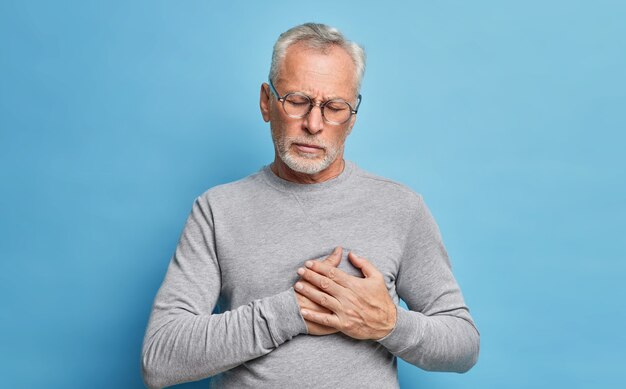 Plan intérieur d'un homme barbu âgé a une crise cardiaque souffre de sentiments douloureux a besoin d'analgésiques appuie les mains sur la poitrine étant malsain porte des lunettes et un pull occasionnel gris isolé sur un mur bleu