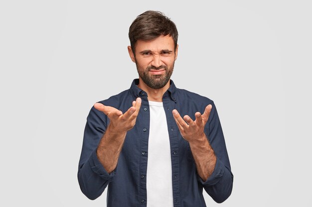 Plan intérieur d'un homme barbu qui fronce les sourcils, a une expression mécontente, des gestes avec les mains, vêtu de vêtements à la mode, isolé sur un mur blanc. Attractive jeune homme européen a l'air bouleversé