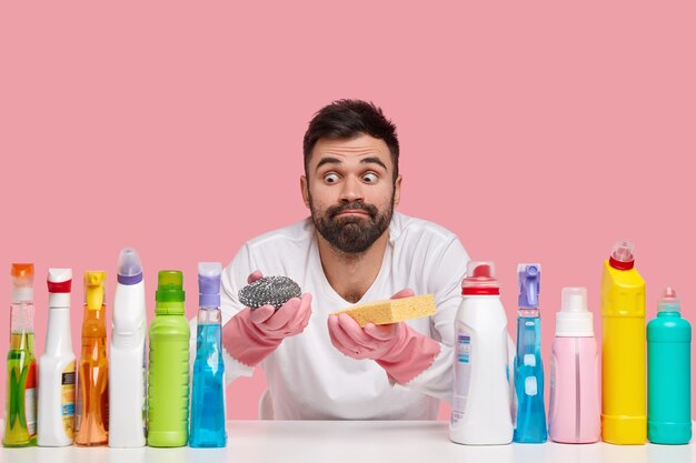 Plan intérieur d'un homme barbu surpris regarde avec les yeux sortis, porte des vêtements décontractés, utilise différents détergents