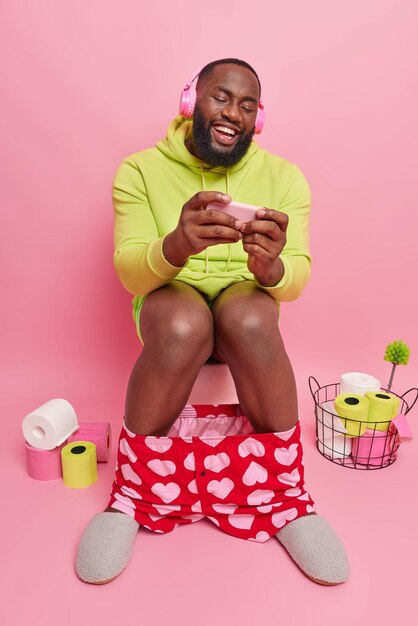 Plan intérieur d'un homme à la peau sombre joue à des jeux vidéo sur un smartphone tout en déféquant sur la cuvette des toilettes porte un sweat à capuche vert utilise des écouteurs stéréo poes dans les toilettes avec des rouleaux de papier autour du concept de dépendance