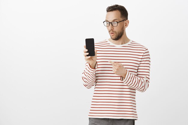 Plan intérieur d'un homme séduisant impressionné étonné dans des verres, montrant un smartphone, pointant et regardant le gadget