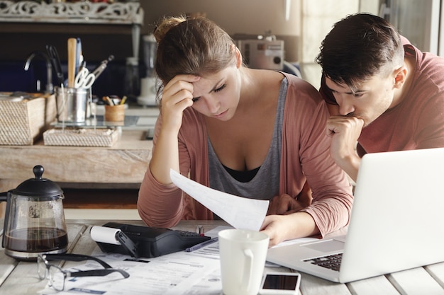 Plan intérieur d'une jeune famille malheureuse en détresse avec des problèmes financiers et des factures de montage, la lecture d'un document avec des regards frustrés tout en calculant les finances domestiques ensemble dans leur cuisine