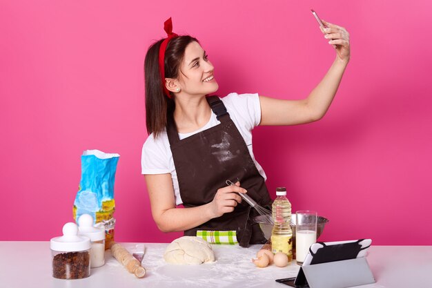 Plan intérieur d'une jeune femme charismatique souriante debout faisant une selfie dans la cuisine tout en préparant un nouveau plat délicieux, en publiant des photos et des vidéos sur les sites de réseaux sociaux. Concept de cuisson et de cuisson.