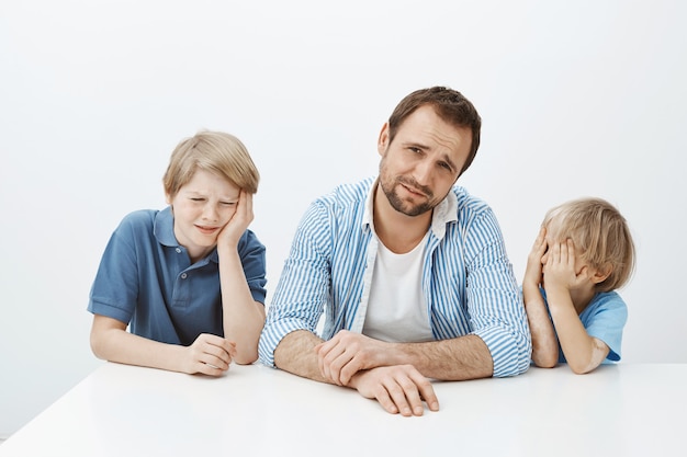 Plan intérieur de jeunes fils mécontents et d'un père assis à table, pleurnichant et bouleversé, fronçant les sourcils de dégoût, voulant manger de la viande pendant que maman donne des légumes