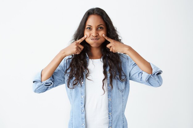 Plan intérieur d'une jolie femme afro-américaine faisant une grimace en soufflant ses joues ayant une expression heureuse. Belle jeune femme à la peau foncée vêtue de vêtements décontractés se moquant.