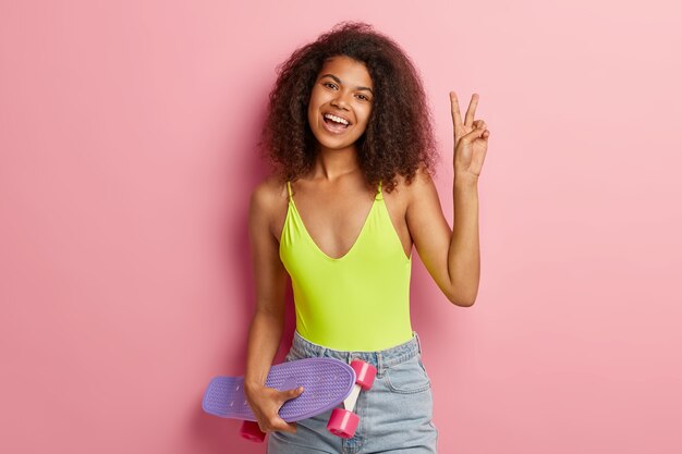 Plan intérieur d'une jolie femme aux cheveux bouclés avec une expression joyeuse, lève la main et fait signe de paix, porte un gilet jaune et un short en jean, a une silhouette mince, tient une planche à roulettes, aime son passe-temps préféré