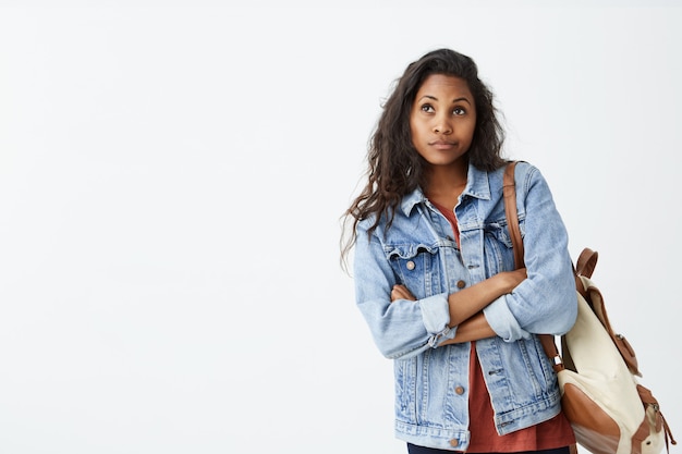 Plan intérieur d'une jolie fille à la peau foncée, détournant les yeux, ayant une expression douteuse et indécise, poursuivant ses lèvres comme si elle n'était pas satisfaite. Jeune femme confuse en veste en jean avec sac à dos