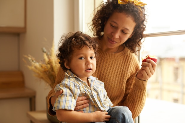 Plan intérieur d'une joyeuse jeune femme portant un pull et un foulard mangeant une pomme sur le rebord de la fenêtre avec un adorable petit garçon potelé sur ses genoux. Liens familiaux, relations, amour et concept parental