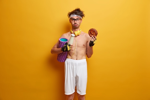 Plan intérieur d'un sportif mécontent ressent la tentation de manger de la malbouffe, porte un rouleau en mousse, veut avoir un corps parfait, porte un casque stéréo autour d'un short, un bandeau, des gants de sport, fait de l'exercice