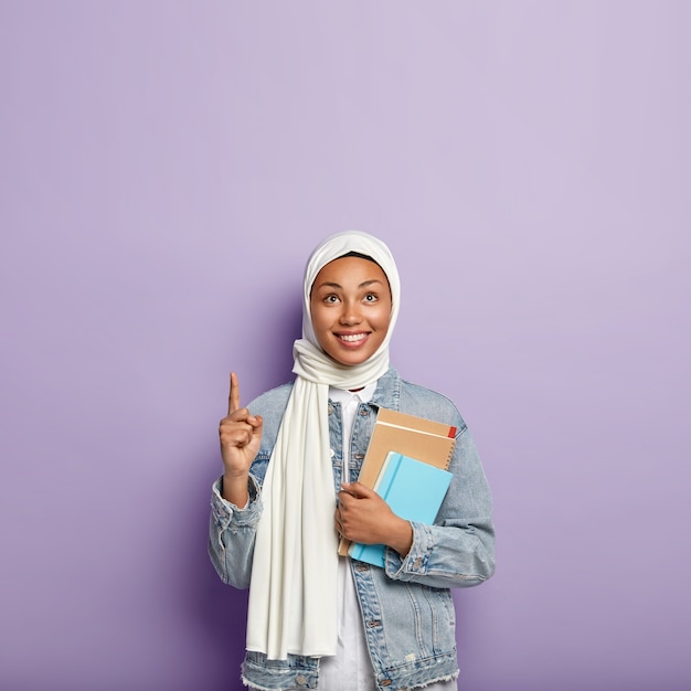 Photo gratuite plan isolé d'une femme religieuse heureuse qui couvre la tête d'un voile, porte un bloc-notes, pointe son index au-dessus, sourit joyeusement, se tient au-dessus d'un mur violet, espace vide pour la promotion. études de fille islamique