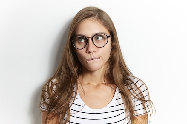 Plan isolé d'une fille européenne hésitante portant des lunettes élégantes, se pinçant les lèvres, ayant une expression faciale perplexe, étant plongée dans ses pensées, pensant à l'examen, regardant ailleurs. Les émotions humaines