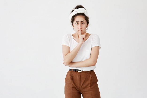 Plan isolé d'une jolie femme brune en colère vêtue d'un t-shirt blanc, ennuyée par un bruit fort, garde l'index sur les lèvres, demande le silence et la paix. Chut, arrête de parler s'il te plait