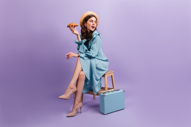 Photo gratuite plan d'une jeune femme en élégante robe bleue. la femme est assise sur un tabouret à côté de la valise et détient un délicieux croissant.