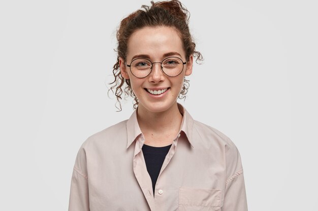 Plan d'une jeune femme européenne positive aux cheveux bouclés foncés, a un sourire doux, une peau tachetée de rousseur, porte une chemise beige décontractée