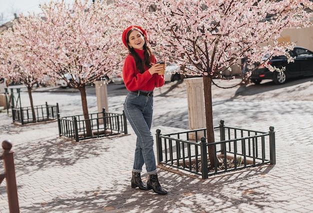 Photo gratuite plan d'une jeune fille en jeans et pull marchant dans le jardin avec un verre de café. teen en béret bénéficiant d'un temps ensoleillé et de boire du thé