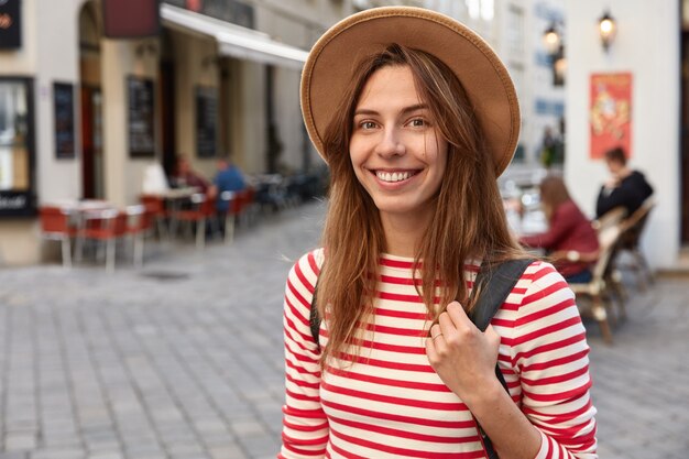 Plan d'une jolie femme sourit joyeusement, porte un chapeau et un pull rayé, étant de bonne humeur en se promenant dans la ville