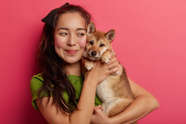 Plan d'une jolie fille coréenne amoureuse de son chien shiba inu, embrasse l'animal avec le sourire, a les cheveux foncés, porte un t-shirt vert, pose avec un animal sur fond rose.