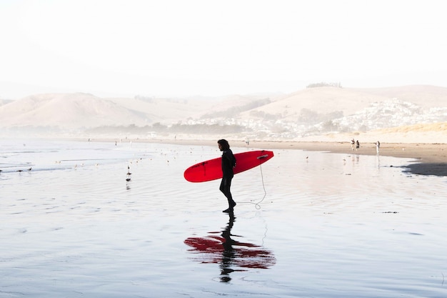 Plan large d'un homme surfeur portant un costume de surf tenant une planche de surf au bord de la mer