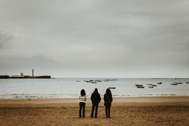 Plan large de trois personnes debout près du bord de mer avec de petits bateaux flottant dans la mer