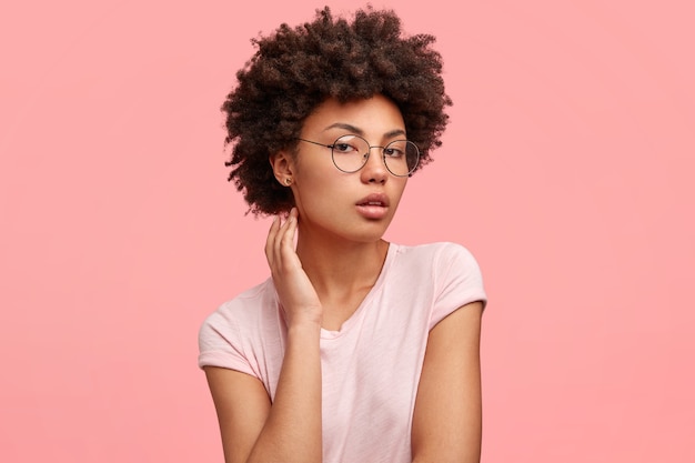 Plan latéral d'une adorable jeune femme à la peau foncée, regarde sérieusement et avec confiance, montre sa beauté naturelle, pose pour un magazine de mode, porte des lunettes rondes et un t-shirt décontracté