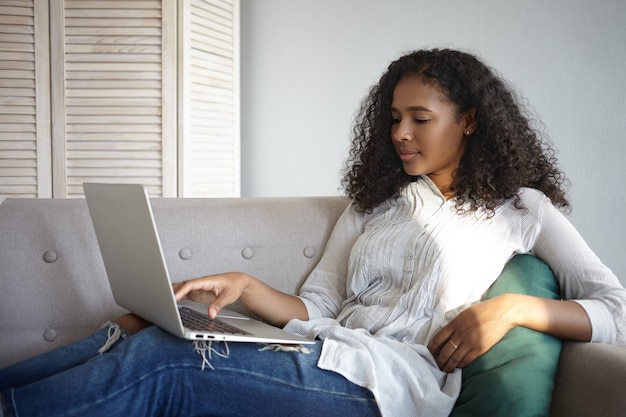 Plan latéral d'une jolie jeune femme afro-américaine regardant un film en ligne sur son ordinateur portable générique tout en se relaxant sur un canapé à la maison seule après le travail. Les gens, la technologie moderne et les loisirs