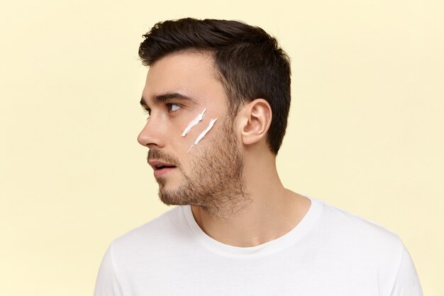 Plan de profil d'un beau jeune homme européen confiant avec une coiffure élégante et des soies appliquant une crème hydratante devant un miroir le matin avant le travail.