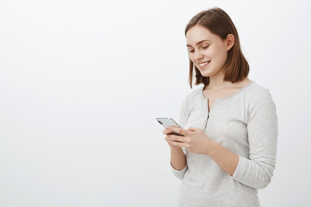 Plan à la taille d'un geek internet féminin insouciant sociable souriant joyeusement tout en ayant une conversation amusante en réseau avec le meilleur ami du jeu en ligne tenant un smartphone et souriant à l'écran du téléphone portable