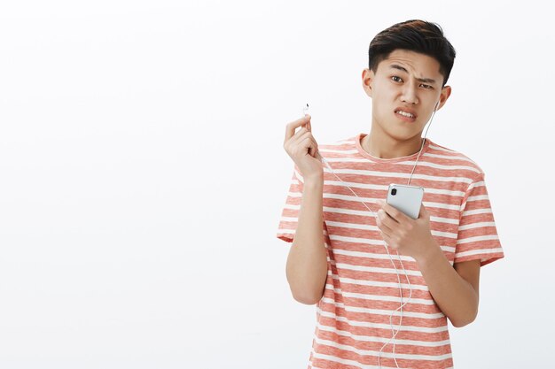 Plan à la taille d'un jeune garçon asiatique mignon mécontent en t-shirt rayé décollant des écouteurs cassés tenant un smartphone fronçant les sourcils insatisfait et dérangé par la qualité du son