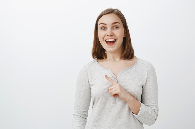 Plan à la taille d'une jolie fille enthousiaste et énergique réagissant à l'offre impressionnante d'un ami allant s'asseoir dans un café souriant joyeusement avec une expression amusée regardant et pointant vers le coin supérieur gauche